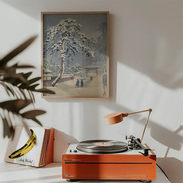Winter landscape painting hanging above a retro record player with an orange base and a lamp in a cozy room setting.
