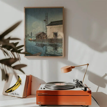 Painting of a tranquil waterfront scene with a traditional building, displayed above a vintage record player in a cozy interior setting.