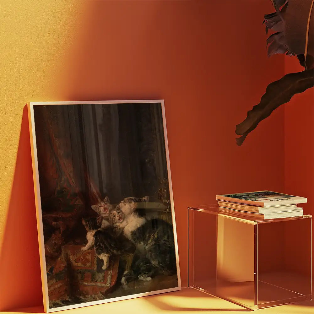 Framed painting of cats resting on a colorful textile, displayed next to a clear acrylic table with stacked books against an orange wall.
