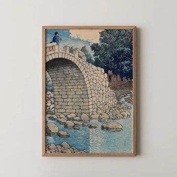 Framed artwork depicting a stone bridge with a person fishing on top, surrounded by a tranquil river and lush greenery.