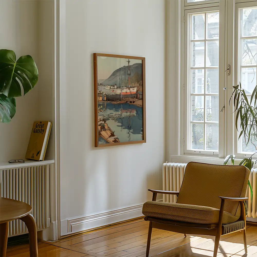 Painting of boats and mountains in a framed art piece displayed on a cream wall, next to a brown chair and a bookshelf with a "Great Gatsby" book.