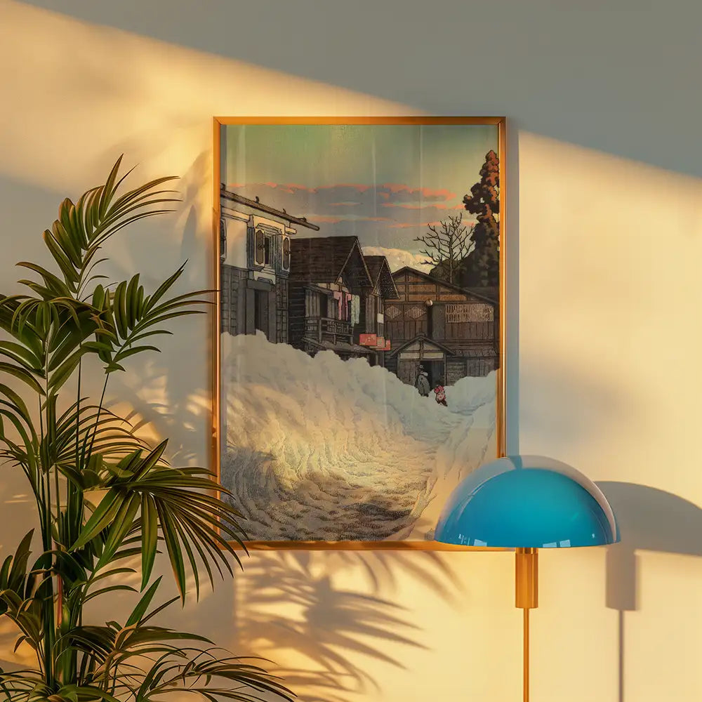 Framed painting of a snowy scene with wooden houses and a sunset, displayed against a wall with a green plant and a stylish lamp.