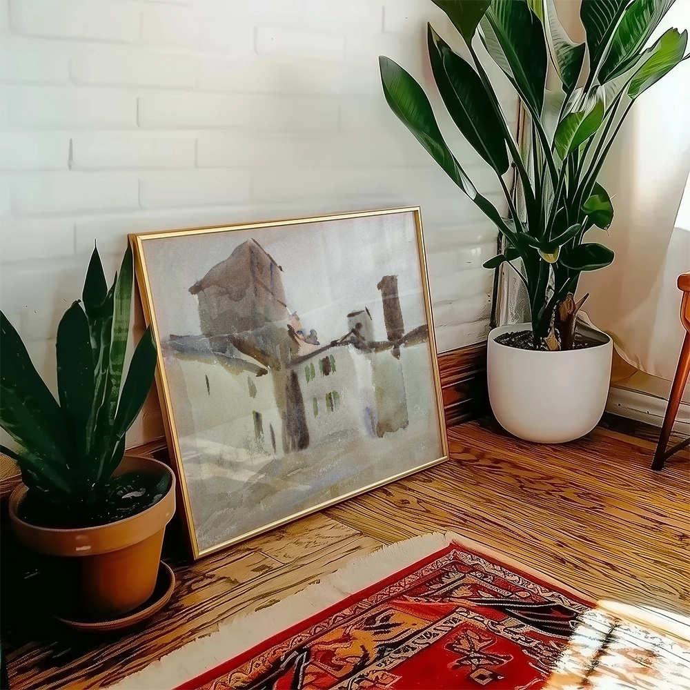 A decorative framed painting of a rustic building placed beside a potted plant in a cozy interior setting.