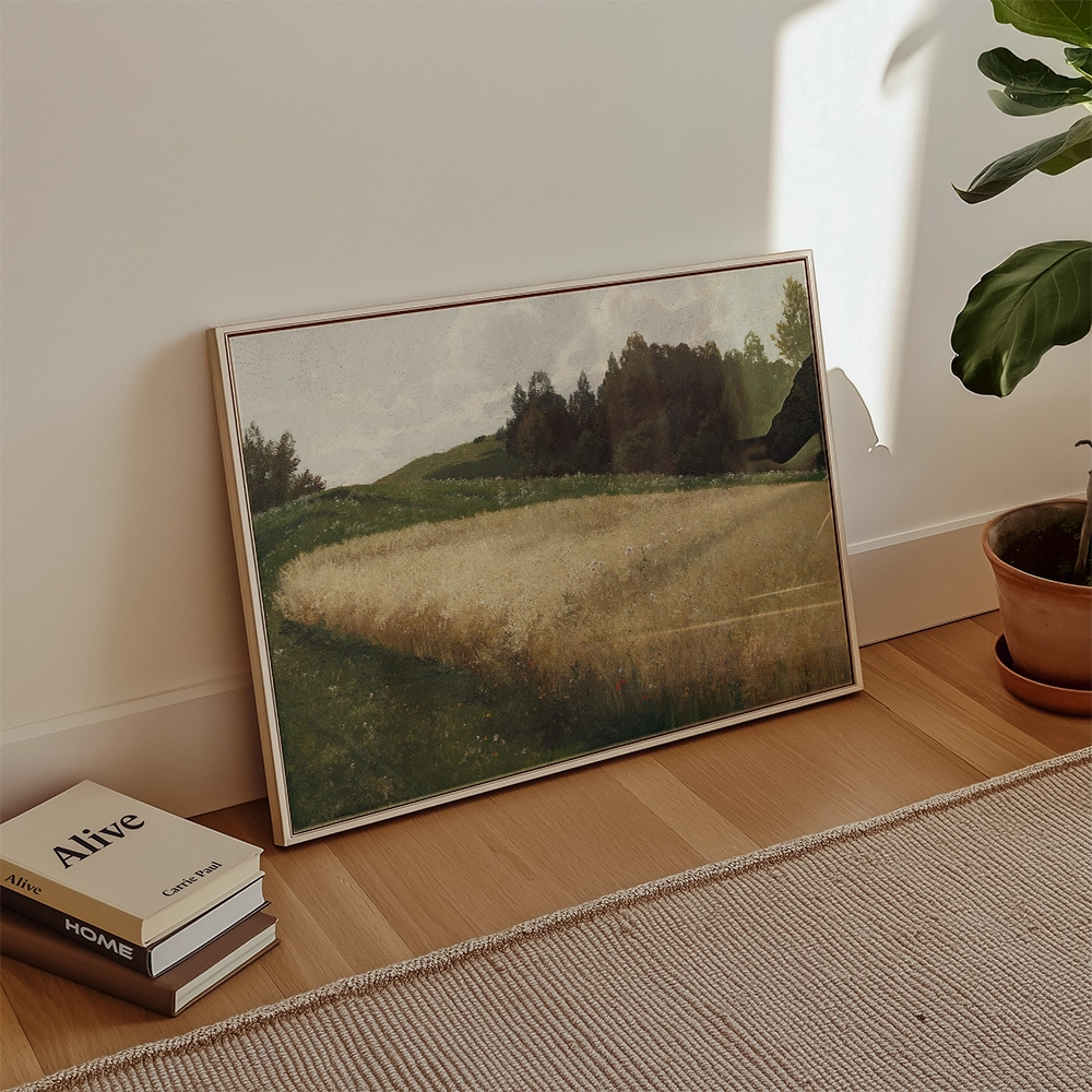 A framed painting of a serene landscape featuring rolling hills and a field of tall grass, leaning against a wall near a potted plant and stacked books.
