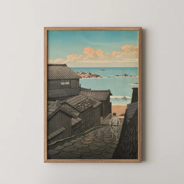 Framed painting of a coastal scene depicting a pathway through traditional buildings leading to the ocean under a blue sky with clouds.