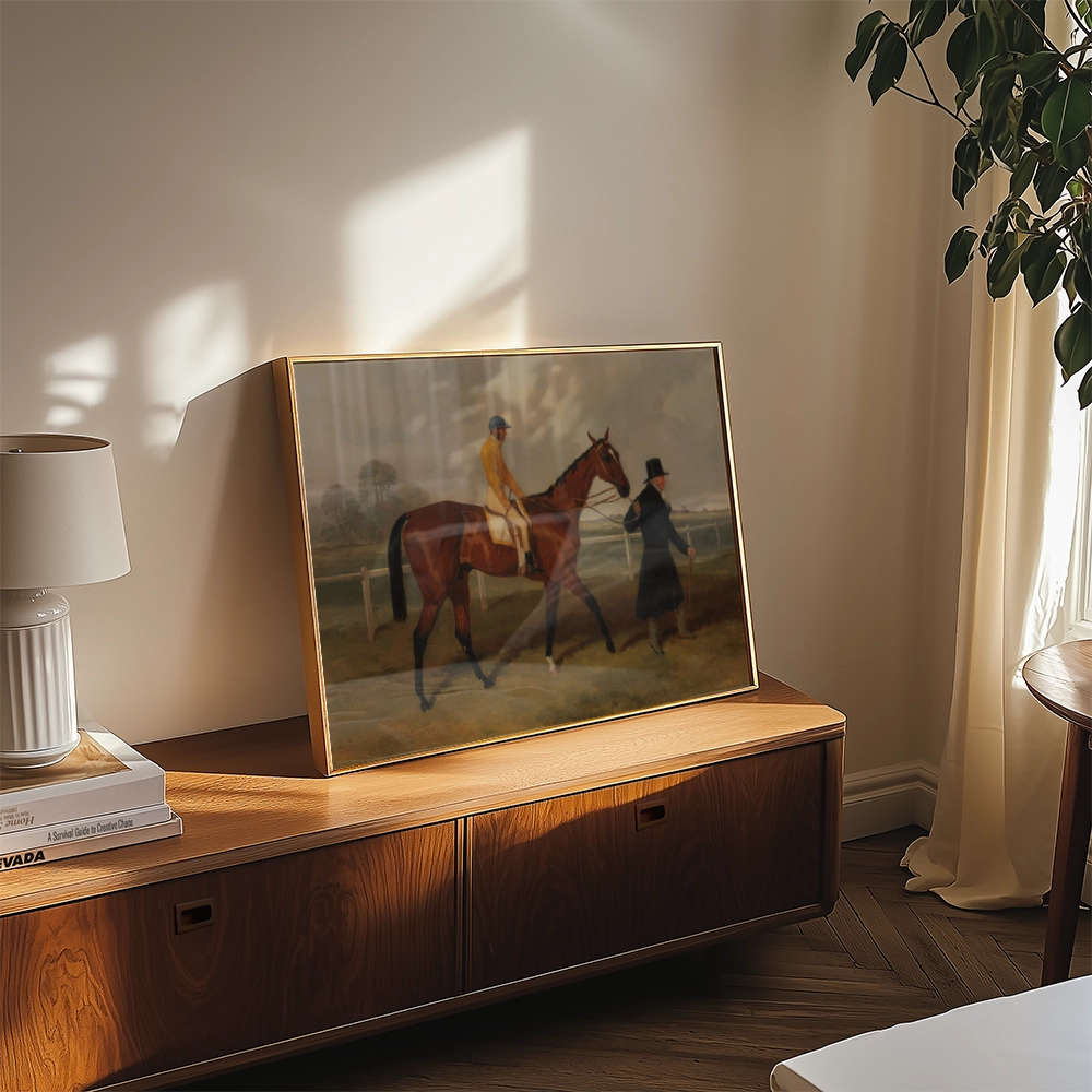 Framed painting of a horse with a rider and a person in a black coat, displayed on a wooden surface in a stylish interior setting.