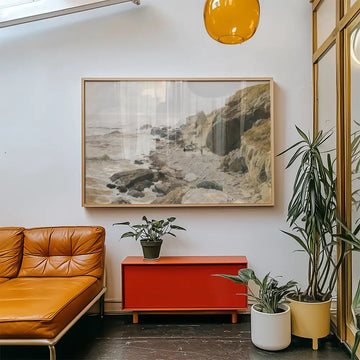 A coastal landscape painting hanging on a white wall above a red cabinet, with a brown leather sofa and green potted plants in the foreground.