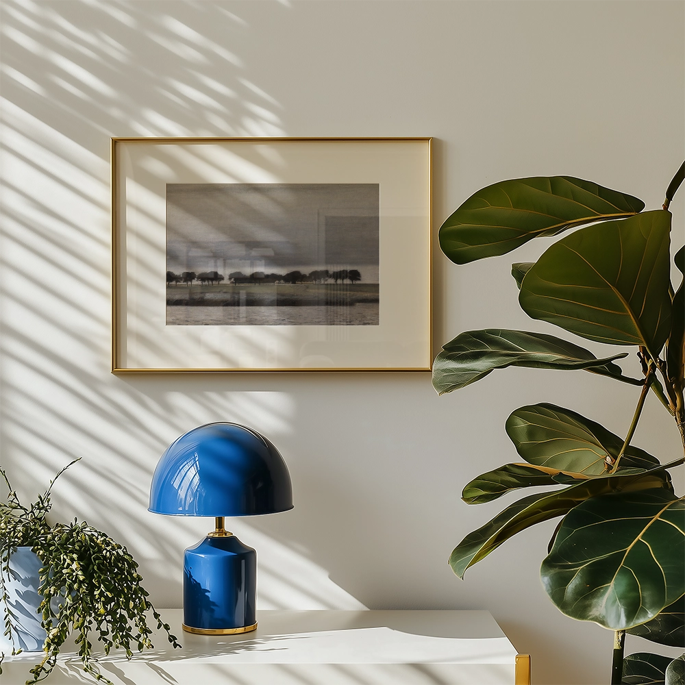 Framed painting of a serene landscape with trees, displayed above a blue lamp and surrounded by indoor plants, on a white table against a light wall.