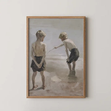 Painting of two boys playing at the beach, one standing and looking on while the other crouches and interacts with the water, framed in a light wood frame.