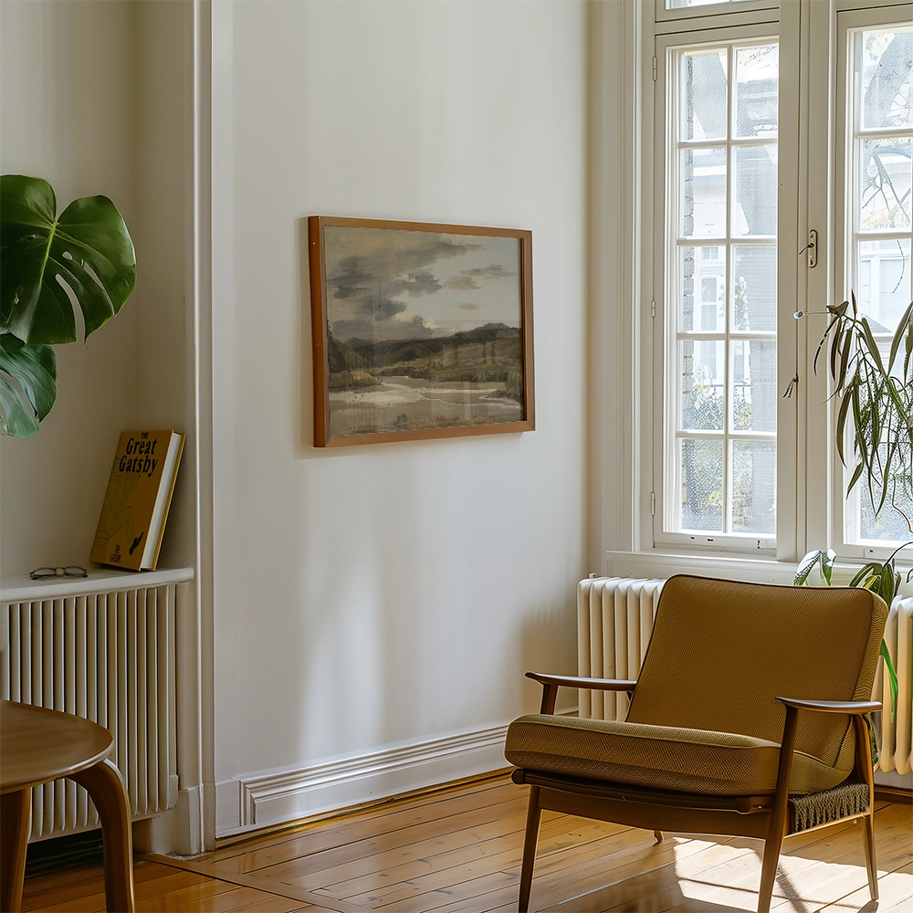 A framed landscape painting hanging on a wall in a bright, modern living room with a plant and a chair.