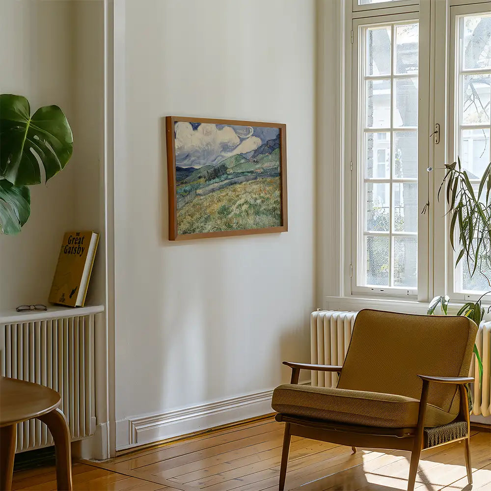 Framed painting of a landscape hanging on a wall in a modern living room with a brown armchair, a green plant, and a book titled "Great Gatsby" on a shelf.