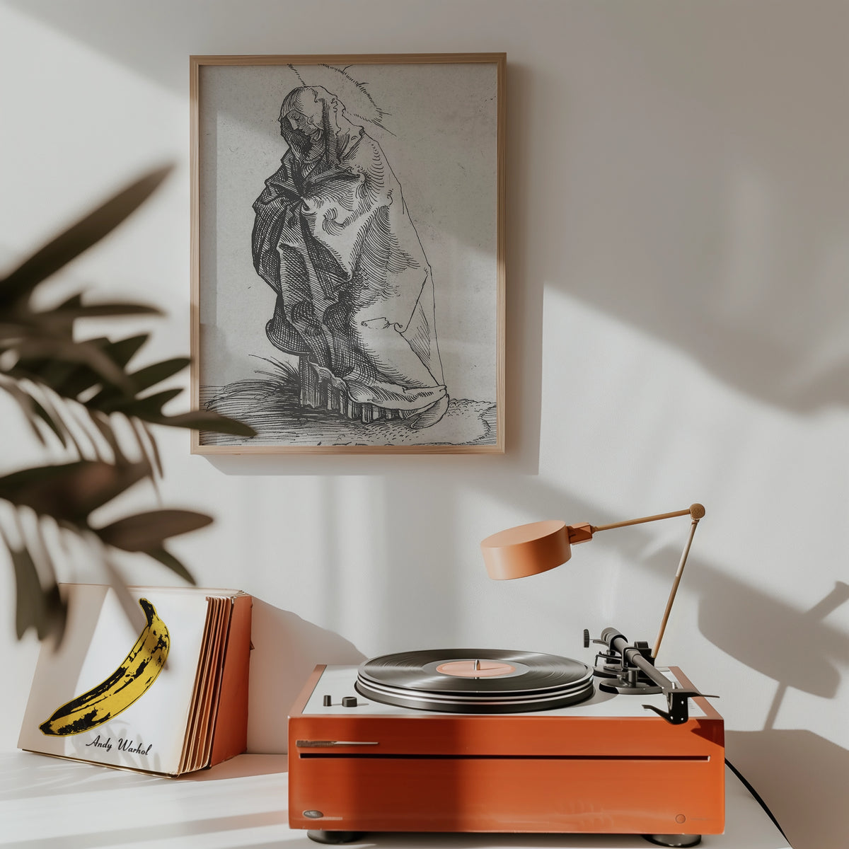 Framed black and white artwork of a cloaked figure on a wall above a stylish orange record player with a lamp and a banana-themed record cover nearby.