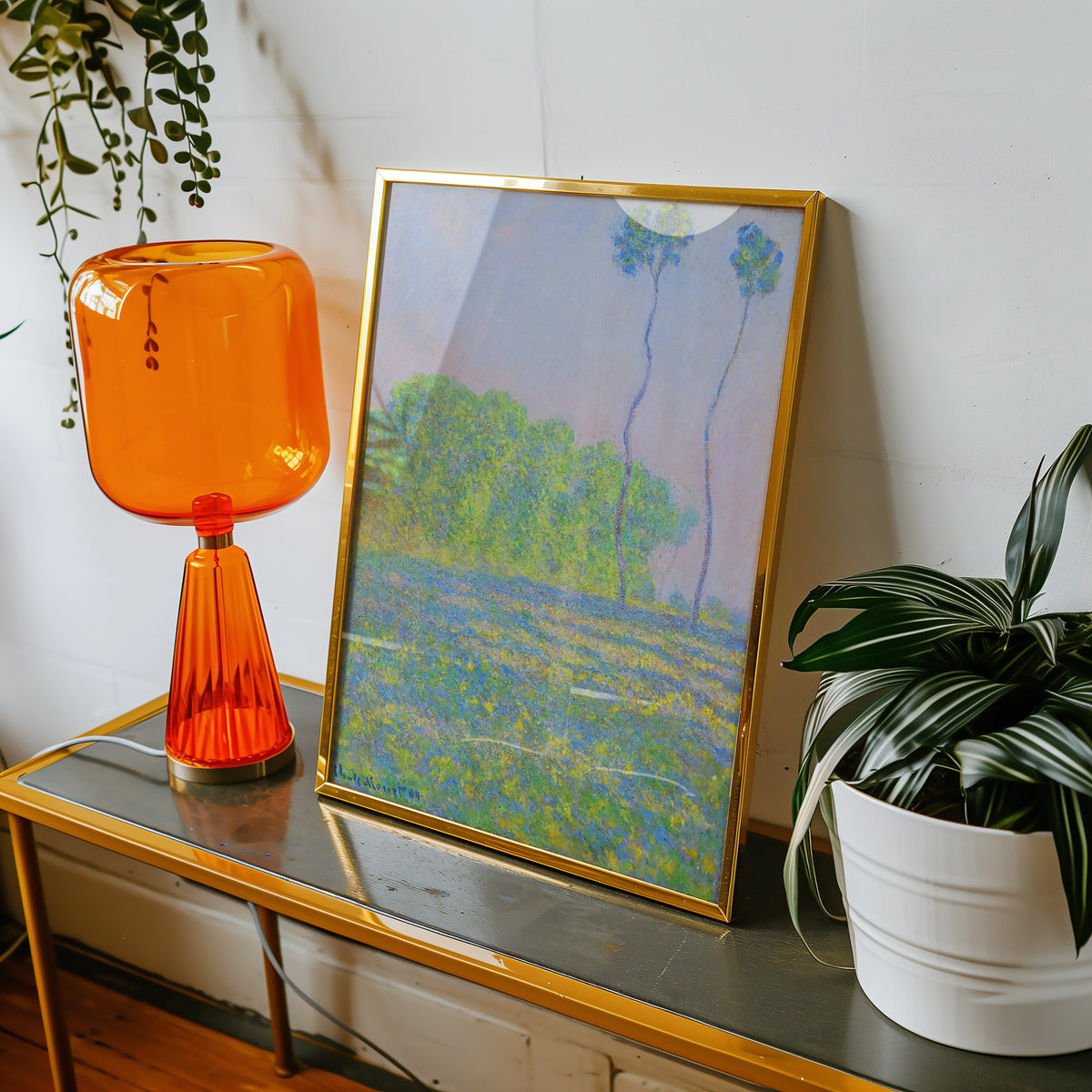 Framed painting featuring a colorful landscape with trees, displayed on a table alongside an orange lamp and a potted plant.