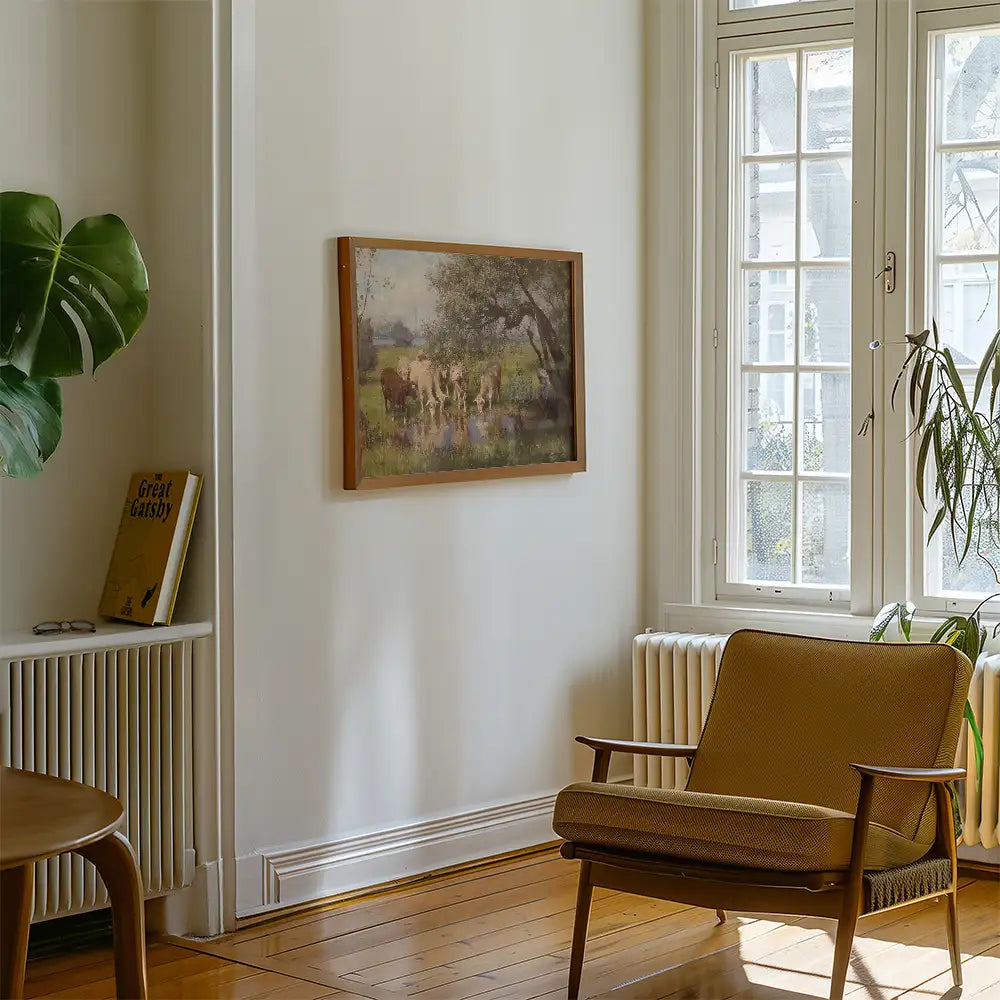 A vintage painting of cows near a water body, framed in wood, displayed on a light wall next to a chair and a plant in a cozy room setting.