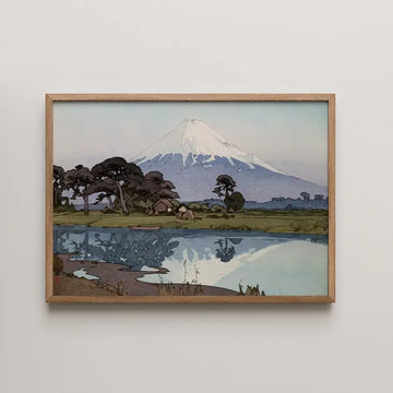 Framed painting of a mountain landscape reflecting in a calm body of water, featuring trees and a small hut in the foreground.