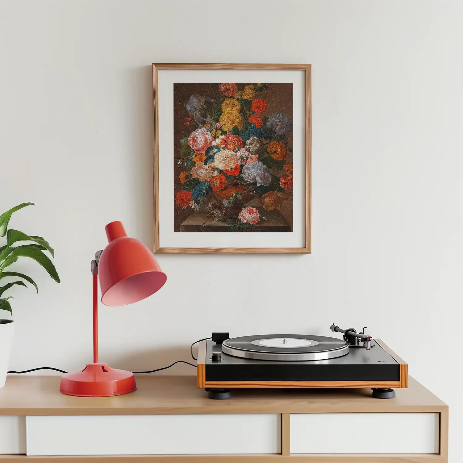 Framed floral painting displayed above a wooden record player and a red desk lamp on a light wood surface.