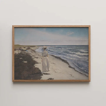 A woman in a white dress and hat walking along a sandy beach with waves crashing on the shore, framed in a wooden frame.