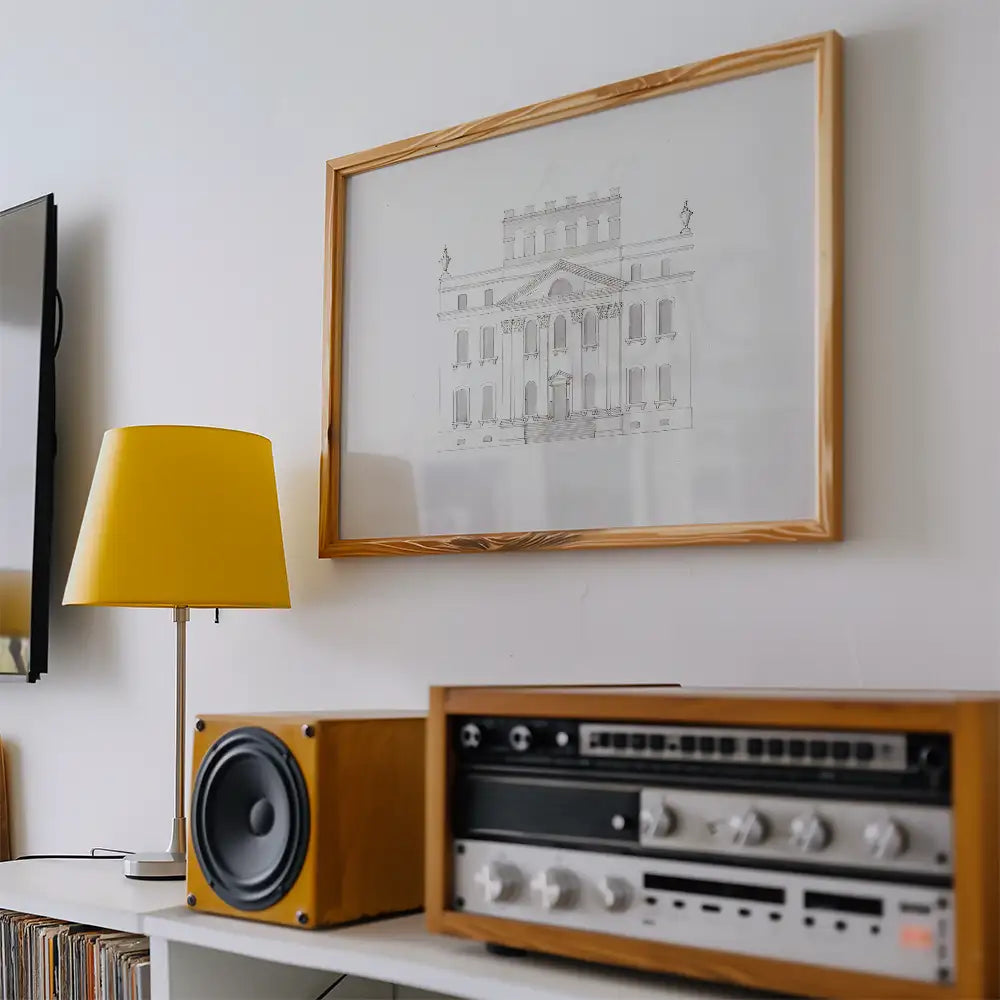 Framed architectural drawing artwork displayed on a white wall, accompanied by a yellow lamp and wooden audio equipment.